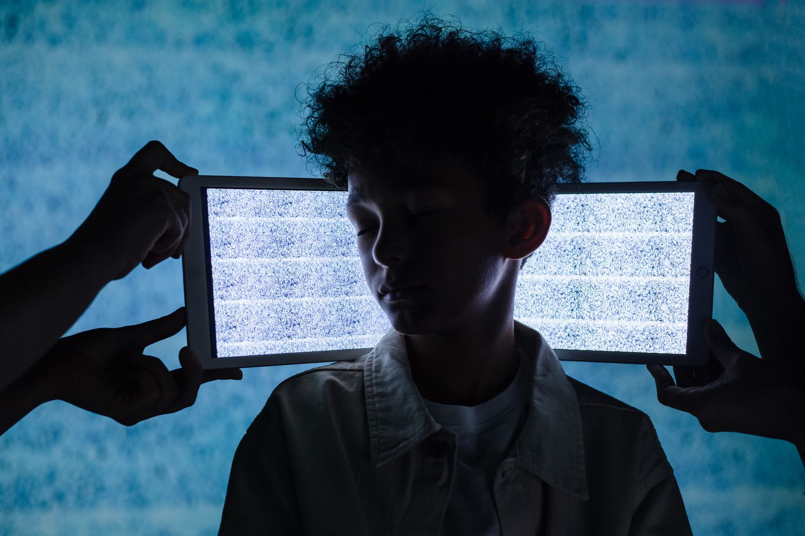 Un adolescent se tient les yeux fermés entre deux tablettes affichant du bruit numérique, symbolisant la surcharge d'inf